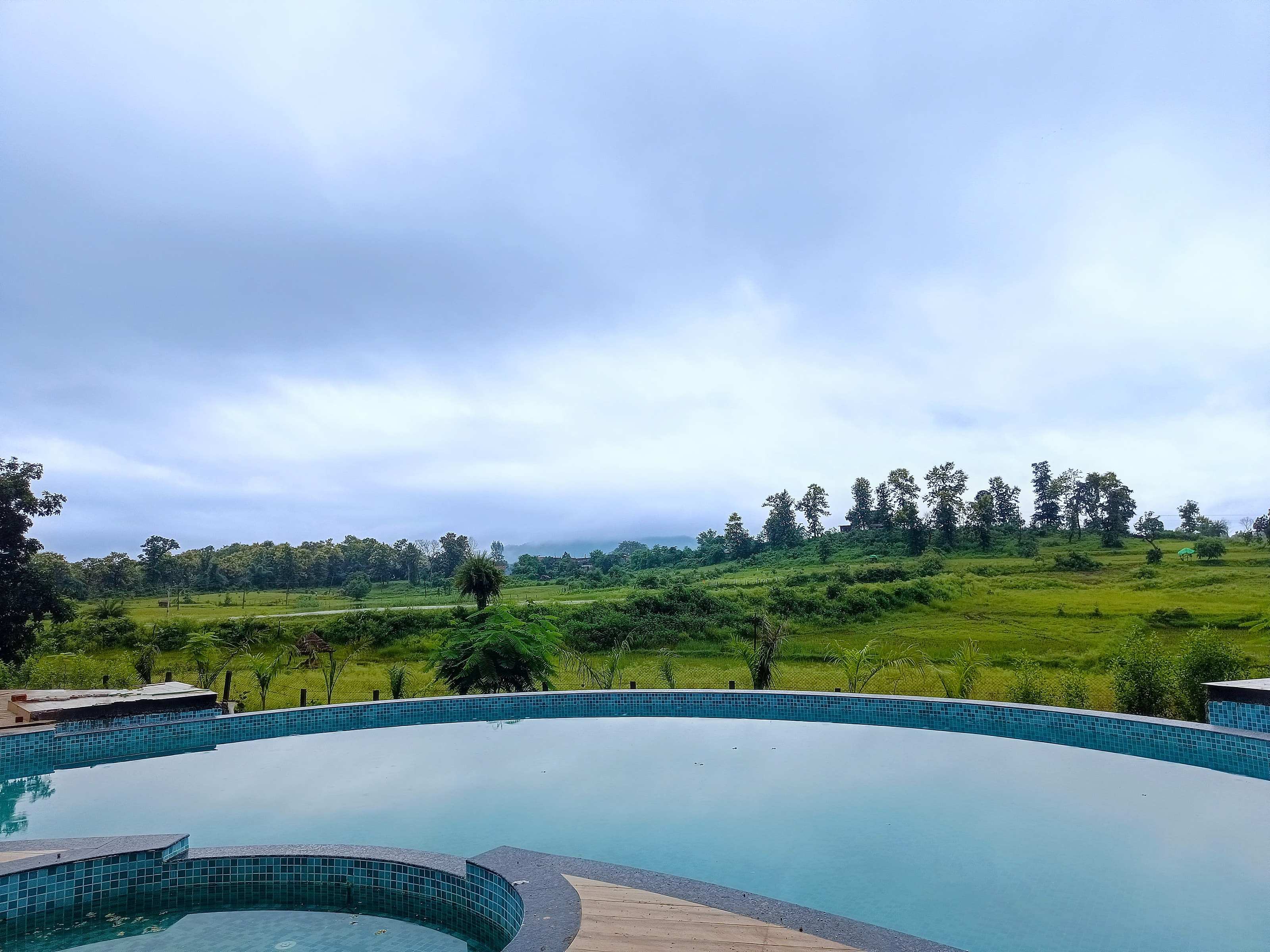 View across the pool edge to open green fields and distant hills under monsoon cloud